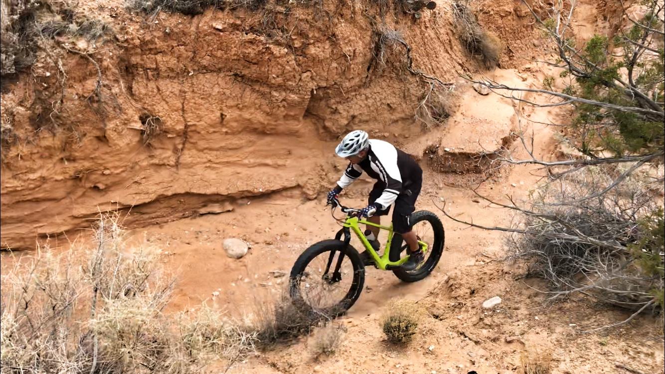 A mountain biker navigating a sandy trail with rocky terrain, wearing a helmet and a black and white jersey. The cyclist is riding a bright green bike, surrounded by dry vegetation and a steep, arid landscape. Mariposa Fat Bike Trails mountain bike trail.