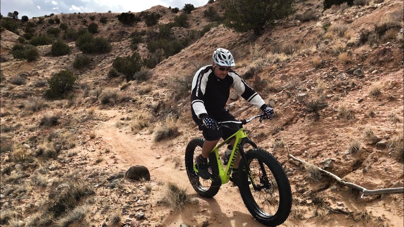 A person riding a fat bike on a dirt trail through a rugged landscape with shrubs and rocky terrain. The cyclist is wearing a helmet and a long-sleeved jersey with gloves, focused on navigating the trail. The scene is set under a partly cloudy sky. Mariposa Fat Bike Trails mountain bike trail.