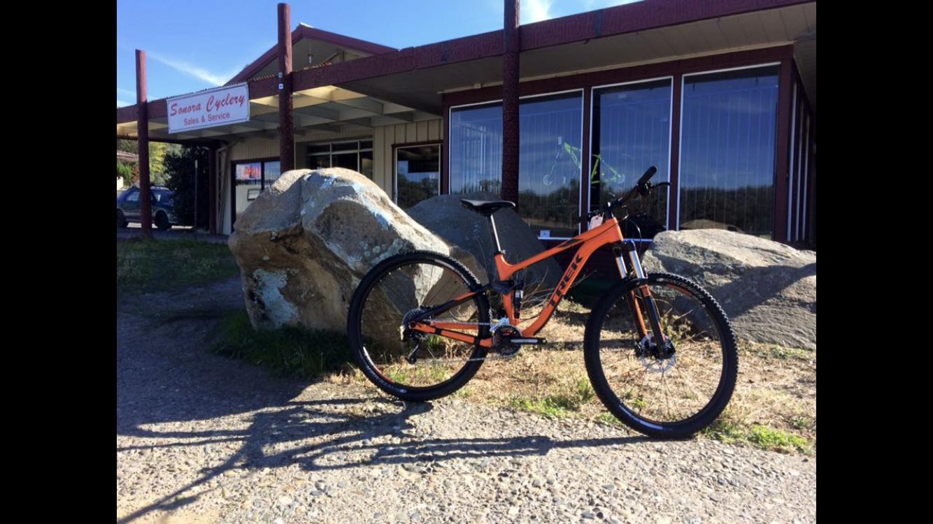 A bright orange mountain bike parked next to a large rock, in front of a cycling shop with a sign reading "Sonora Cycling Sales & Service." The shop features large windows and is surrounded by grassy terrain on a sunny day.