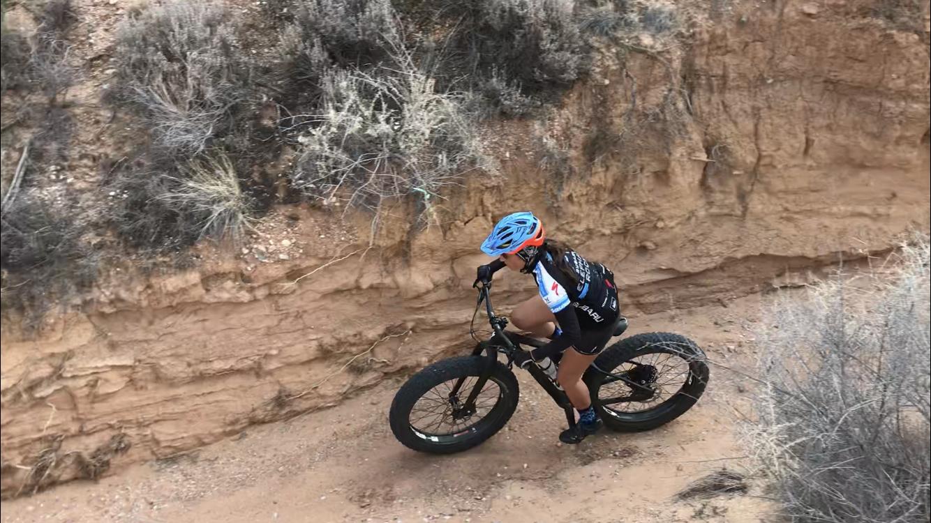 A cyclist riding a fat bike on a dirt trail in a rugged outdoor setting, surrounded by dry shrubs and sand-colored terrain. The cyclist is wearing a blue helmet and a black jersey with colorful accents, focused on navigating the steep, uneven path. Mariposa Fat Bike Trails mountain bike trail.