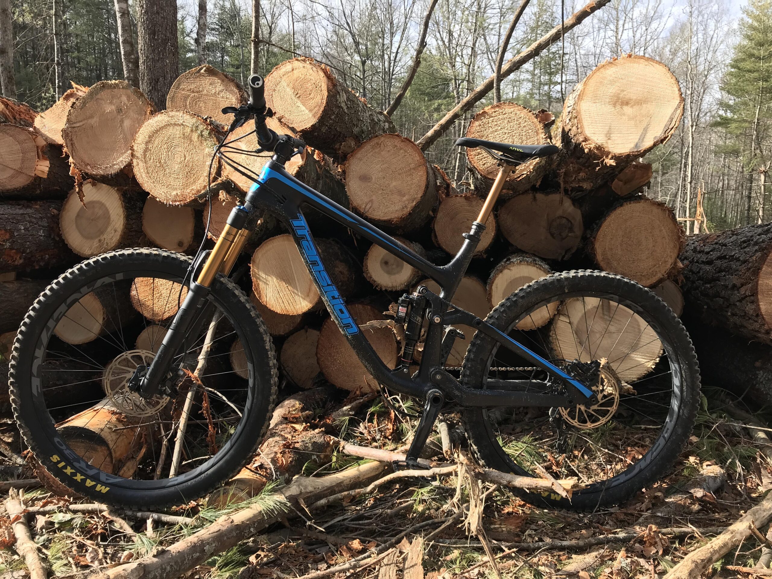 Transition Patrol: A black and blue mountain bike is positioned against a stack of freshly cut logs in a forest setting, with trees in the background. The bike features prominent suspension forks and is resting on the forest floor covered in fallen branches and pine needles.