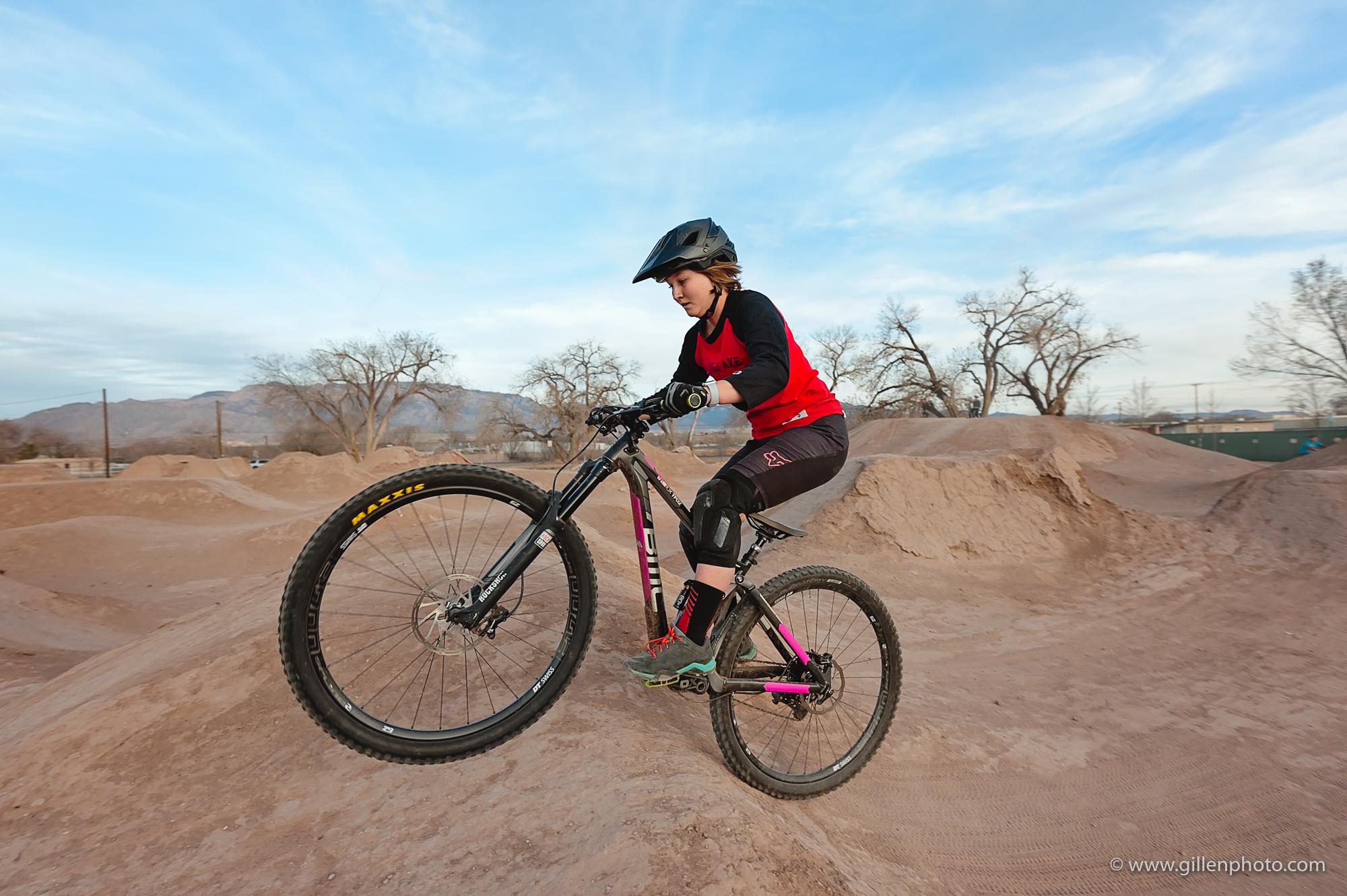 A young rider in a red and black shirt is performing a jump on a mountain bike at a dirt biking park, with a background of sandy terrain and sparse trees under a blue sky. The rider is wearing a helmet and protective gear while biking on the uneven surface. North Valley Bike Park mountain bike trail.