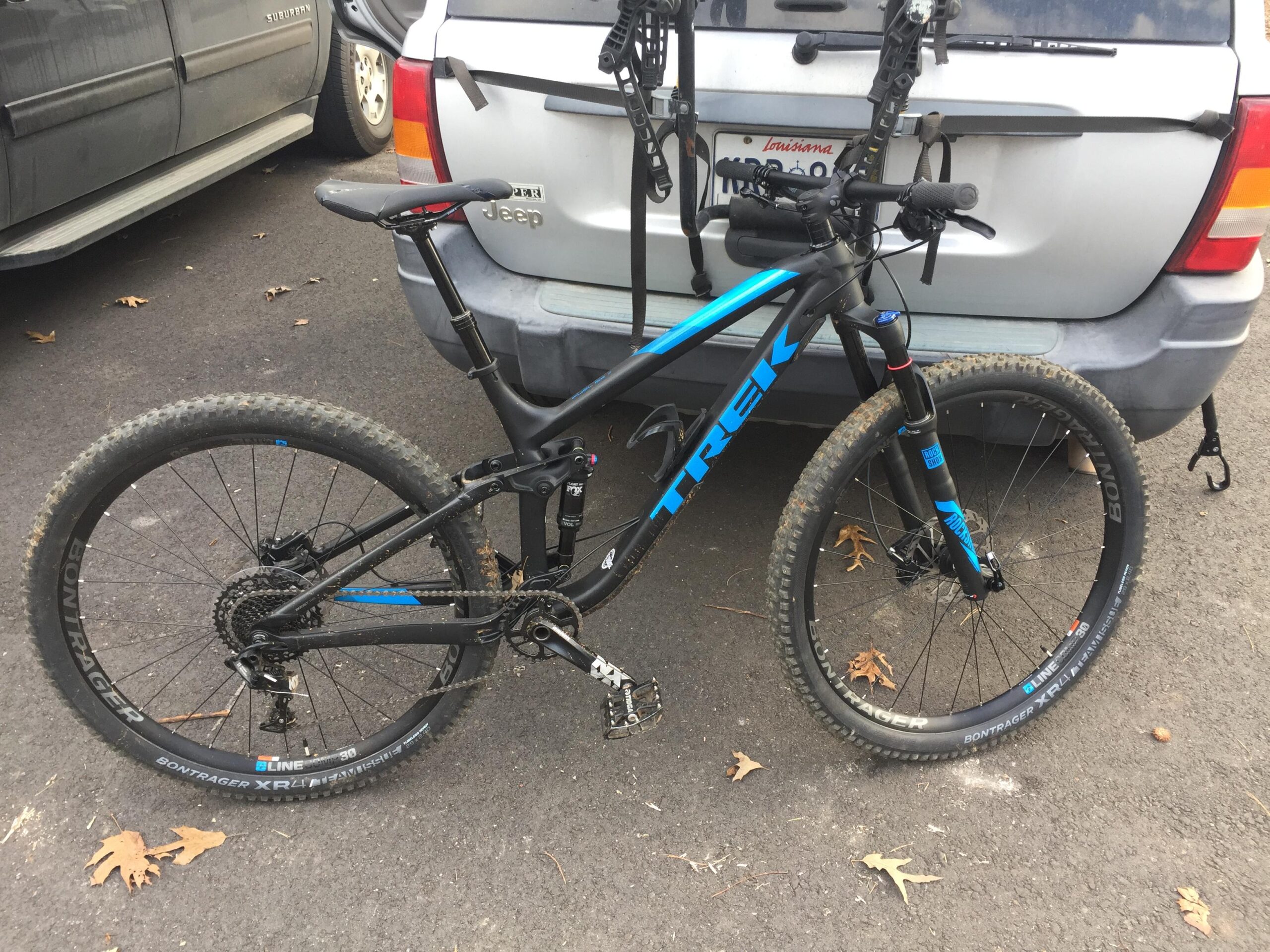 Trek Fuel EX 7 29: Alt text: A black and blue Trek mountain bike is parked beside a silver SUV with a Jeep emblem. The bike features knobby tires and has some mud on the wheels, indicating recent use.