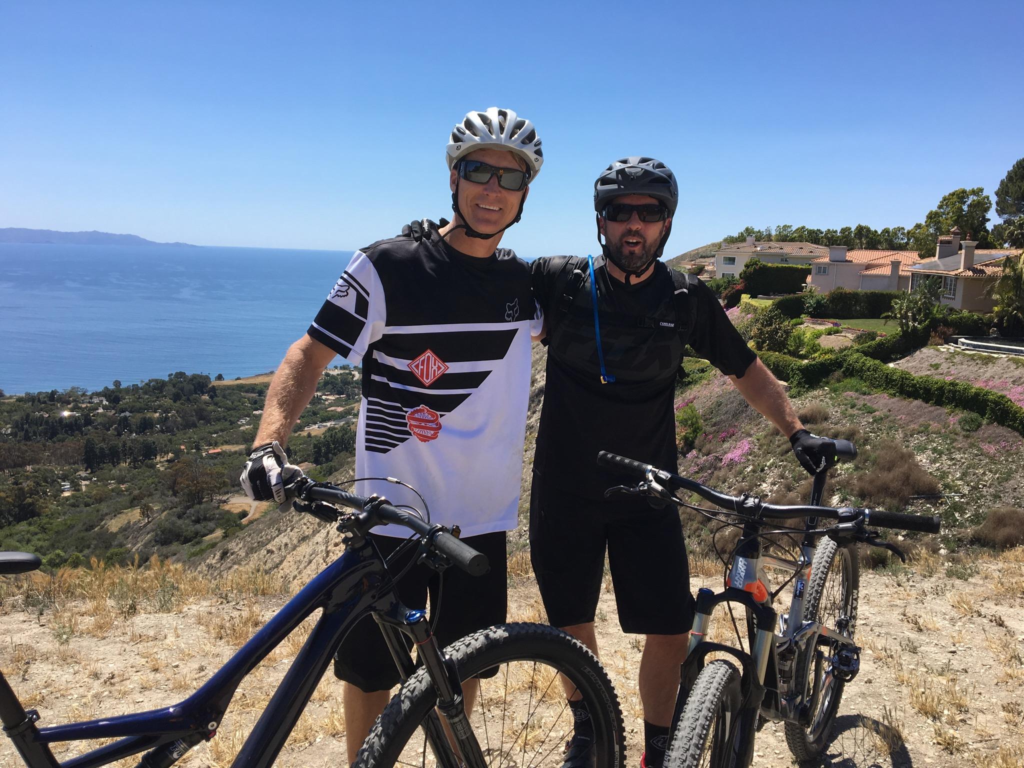 Specialized Stumpjumper FSR comp carbon 650B: Two mountain bikers pose together on a hillside overlooking the ocean. They are wearing helmets and sunglasses, dressed in athletic gear. The background features a scenic view of the coastline and residential homes. The landscape is rugged with dry grass and blooming flowers.