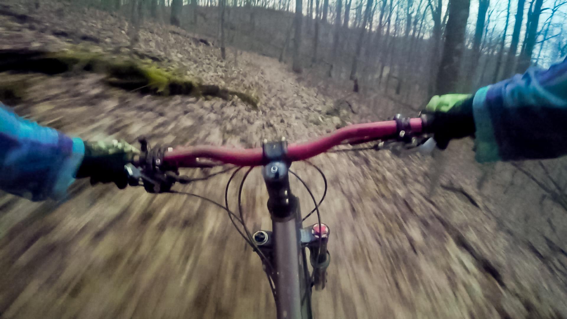 A first-person perspective of mountain biking on a forest trail, with hands gripping a red bike handlebar. The ground appears blurred, suggesting high speed as trees and nature surround the path. Kanawha State Forest Trails mountain bike trail.