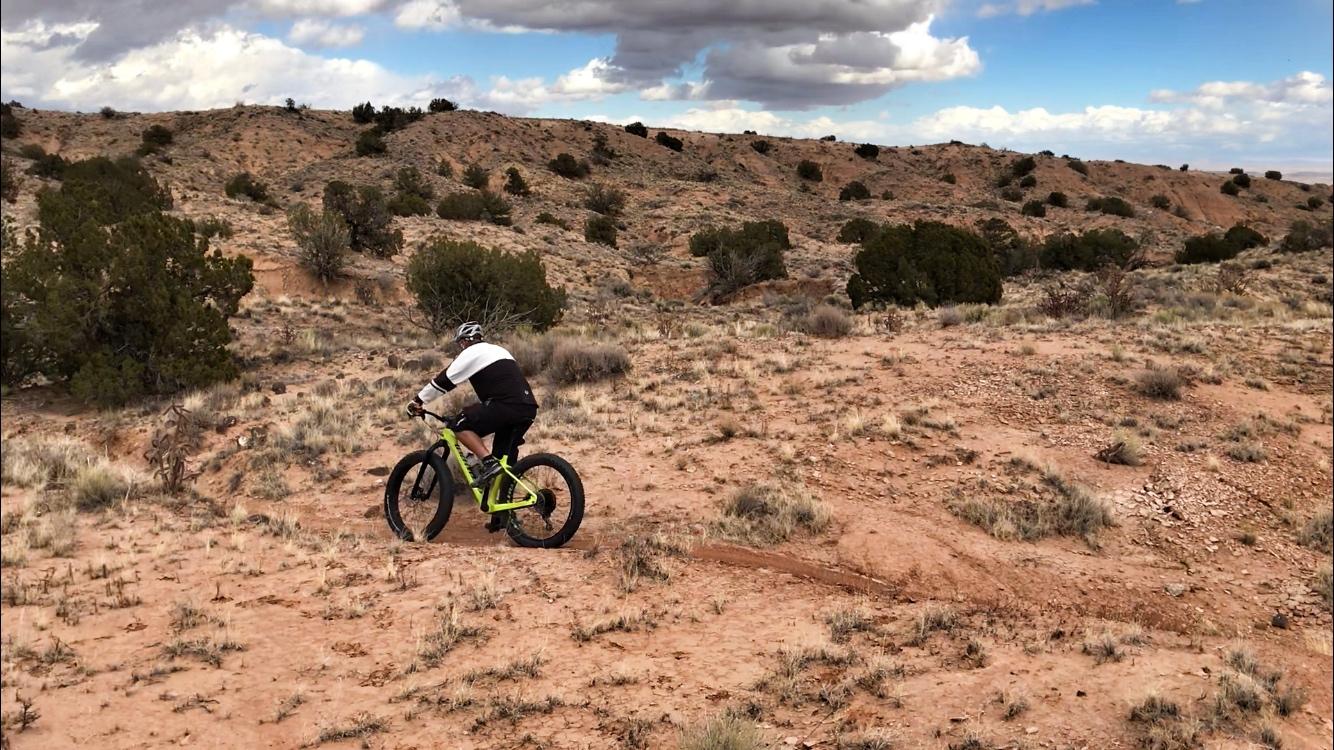 A person riding a mountain bike on a dirt trail in a rugged, arid landscape with sparse vegetation and rocky terrain, under a partly cloudy sky. Mariposa Fat Bike Trails mountain bike trail.