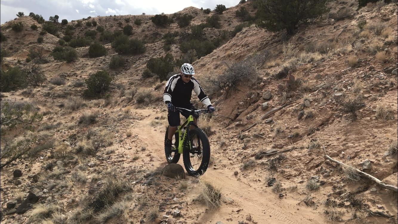 A person riding a fat bike on a dirt trail through a rocky, arid landscape. The cyclist is wearing a helmet and cycling gear, navigating over a sizable rock on the path, with shrubs and small trees visible in the background. The sky is partly cloudy, suggesting an outdoor adventure. Mariposa Fat Bike Trails mountain bike trail.