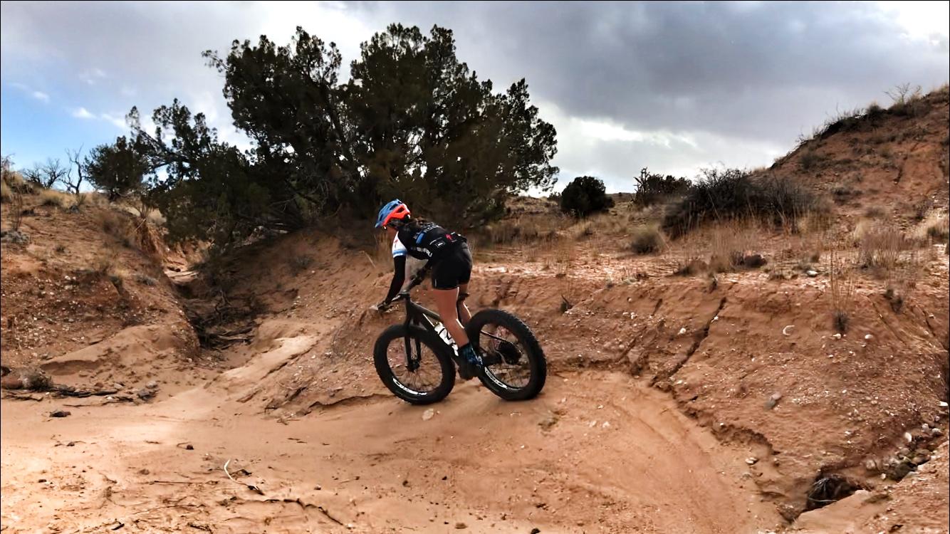 A person riding a fat tire mountain bike along a sandy, uneven trail surrounded by sparse vegetation and trees under a cloudy sky. Mariposa Fat Bike Trails mountain bike trail.
