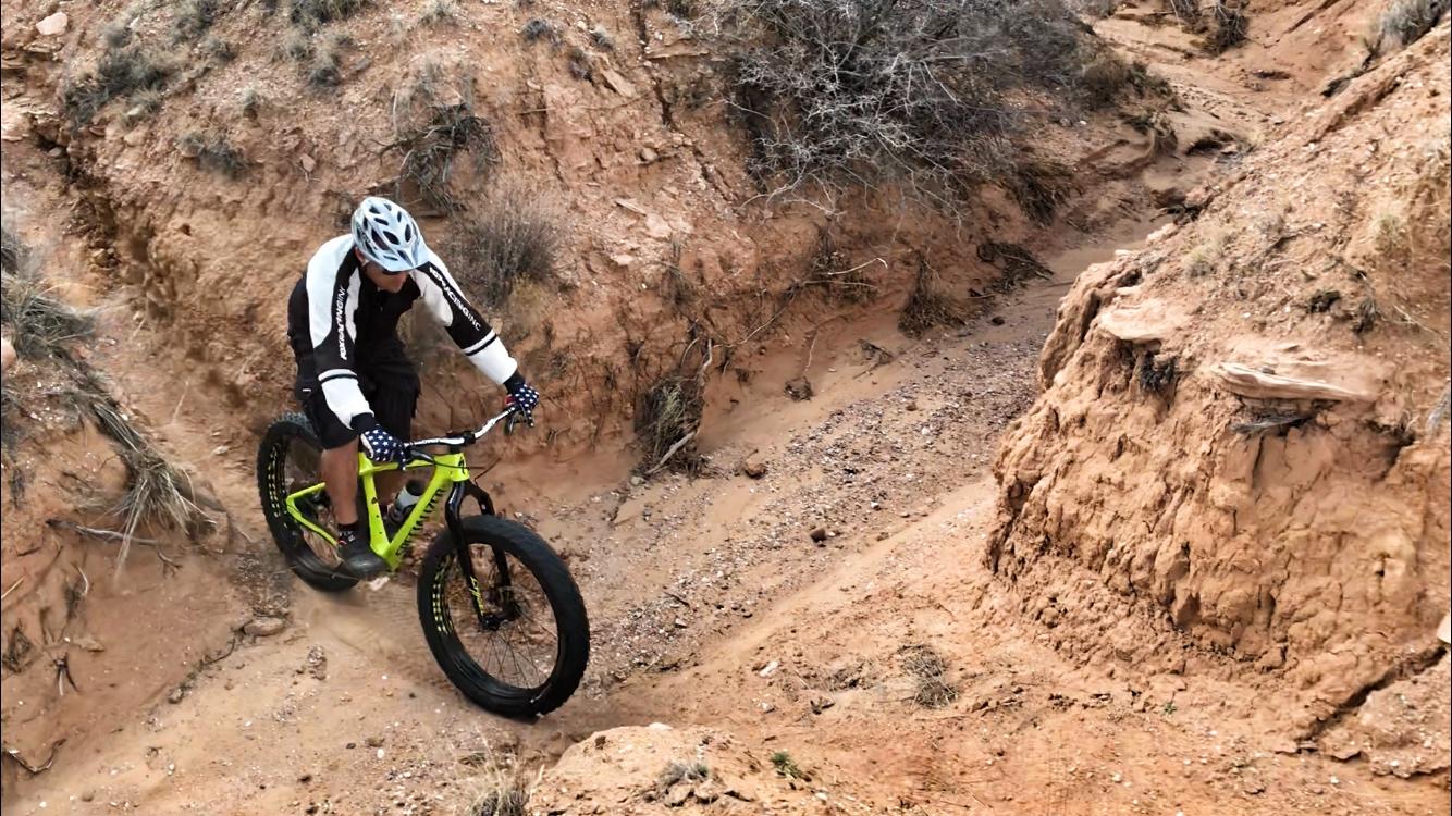 A cyclist riding a bright green mountain bike down a sandy, narrow trail surrounded by rocky terrain and sparse vegetation. The rider is wearing a helmet and a black and white cycling outfit, focusing on navigating the uneven path. Mariposa Fat Bike Trails mountain bike trail.
