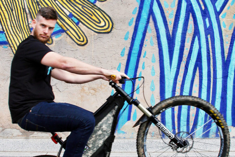 A person performing a wheelie on a mountain bike in front of a colorful mural featuring blue abstract designs. The individual has short hair and a beard, wearing a black t-shirt and blue jeans, with a focused expression. The bike has a camouflage bag attached to its frame.