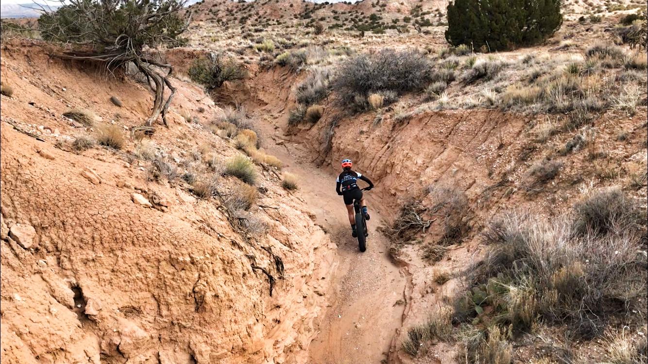 A mountain biker navigates a narrow dirt trail surrounded by arid terrain, with sparse vegetation and rocky hills in the background. Mariposa Fat Bike Trails mountain bike trail.