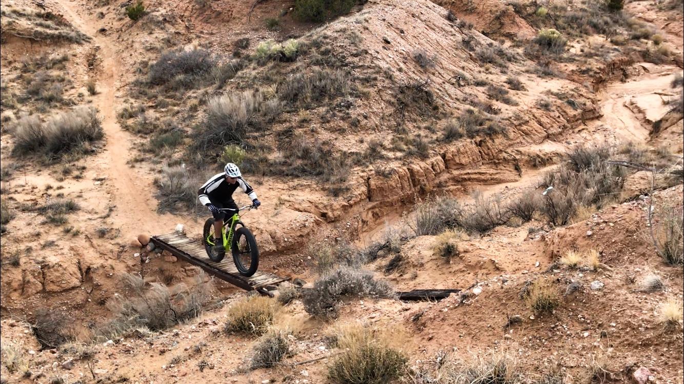 A mountain biker wearing a black and white jersey and helmet rides over a narrow wooden bridge on a rugged trail surrounded by dry vegetation and rocky terrain. Mariposa Fat Bike Trails mountain bike trail.