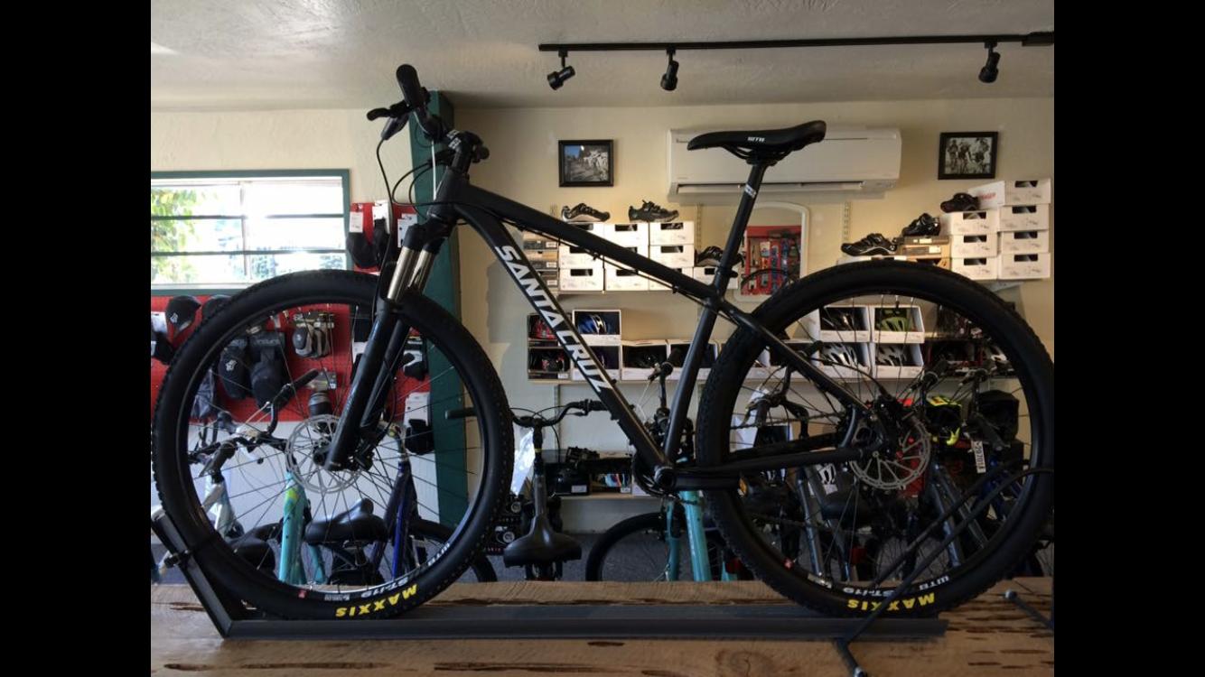 A black mountain bike displayed in a bicycle shop, featuring a sleek design and the brand name "Santa Cruz" prominently on the frame. The bike is positioned on a wooden platform, with various biking accessories and equipment visible in the background.
