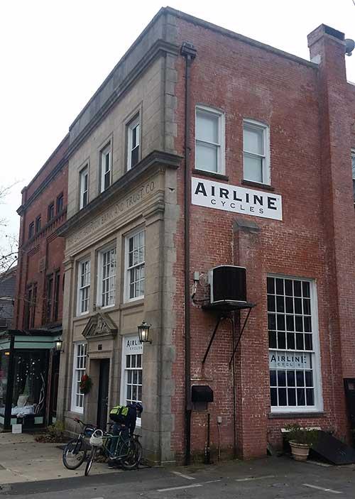 A brick building with an old architectural style featuring a stone façade labeled "BUILDING TRUST CO" and a modern sign reading "AIRLINE CYCLES." The corner of the building shows large windows and an air conditioning unit, while a bicycle is parked in front. Nearby, there is a glass storefront with a small awning. The photo captures a quiet street scene, likely in an urban area.