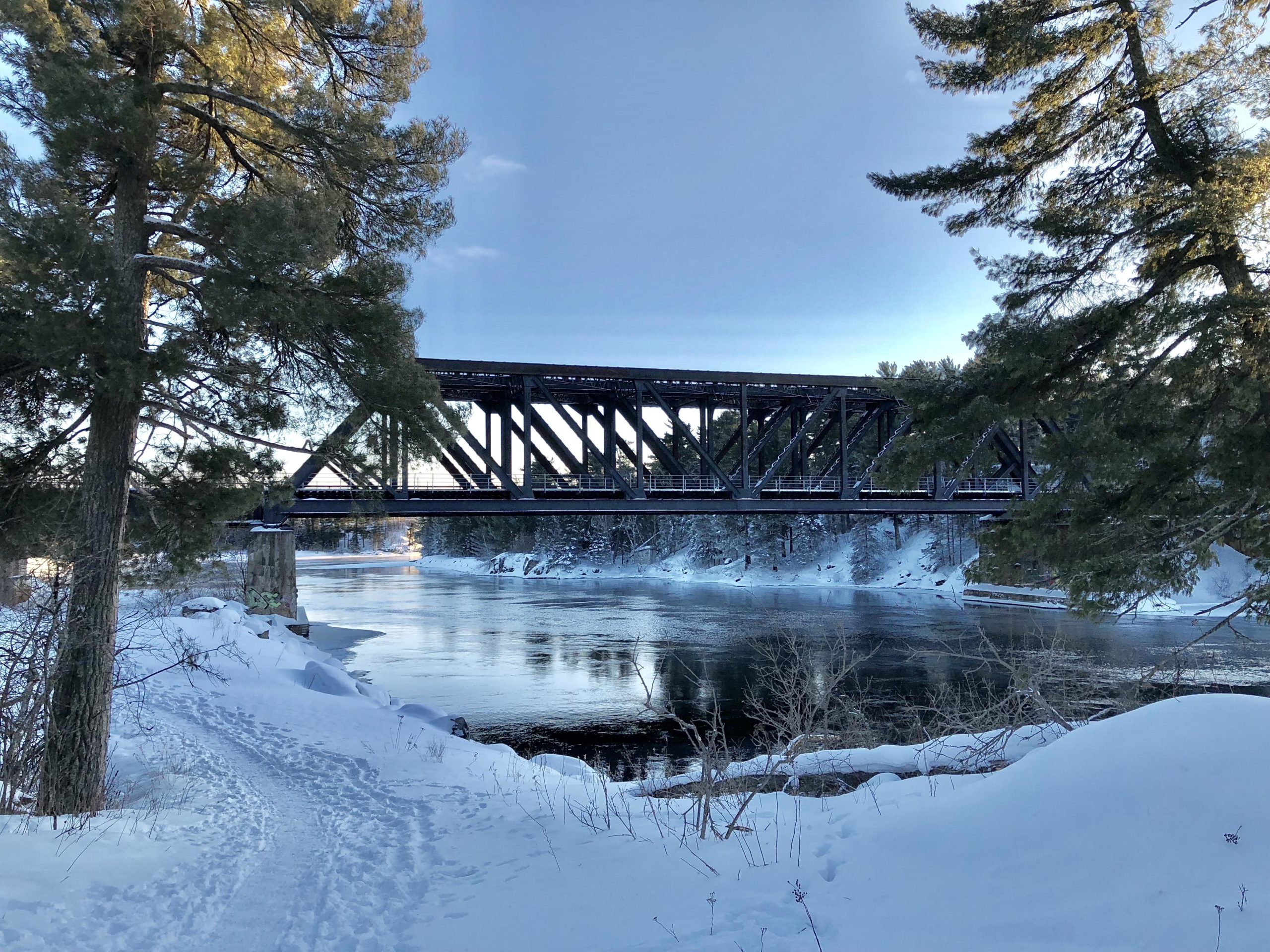 A snowy landscape featuring a metal railway bridge spanning a river, surrounded by tall pine trees. Snow-covered ground and a clear blue sky create a tranquil winter scene. Tunnel Trails mountain bike trail.