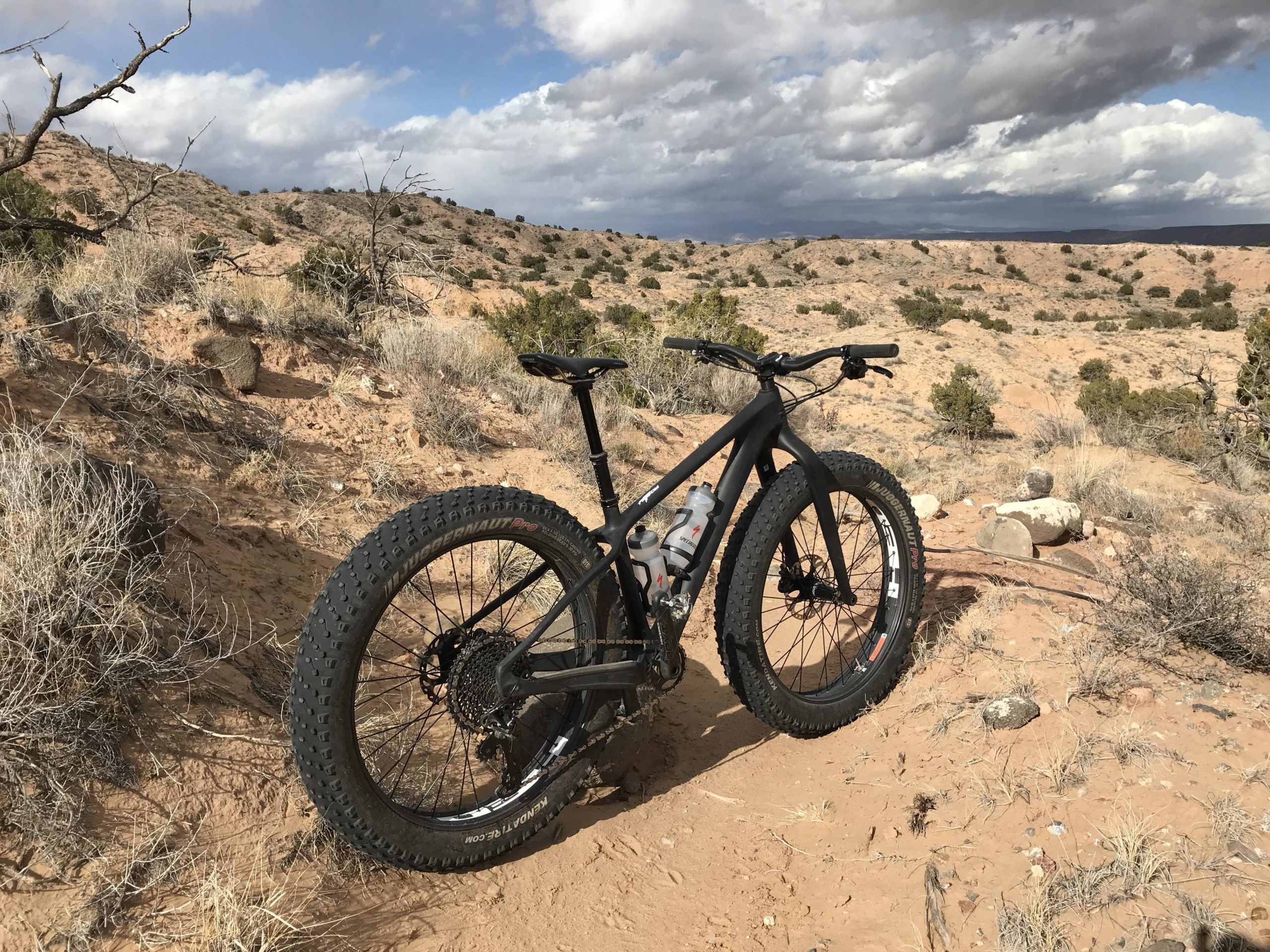 A black fat bike parked on a sandy trail in a rugged landscape, surrounded by sparse vegetation and rocky terrain under a partly cloudy sky. Mariposa Fat Bike Trails mountain bike trail.