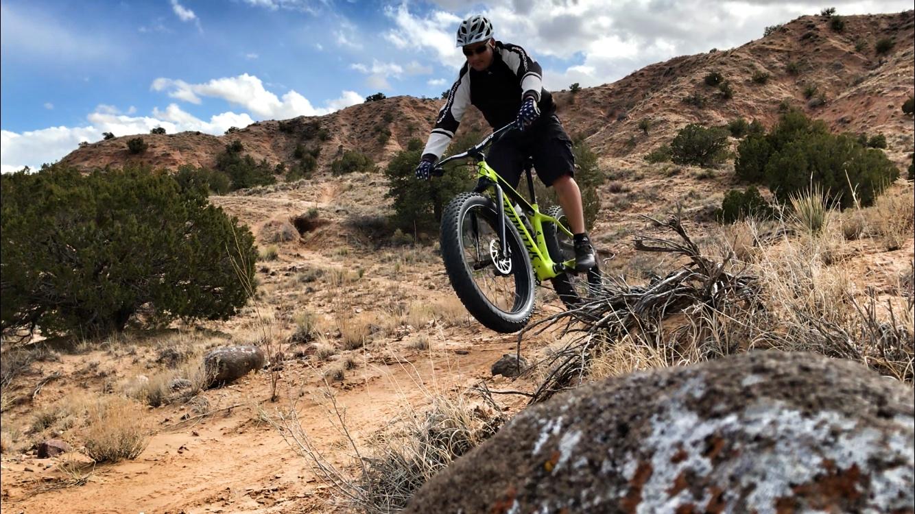 A person riding a fat bike performs a jump on a rocky dirt trail surrounded by shrubs and hills under a partly cloudy sky. Mariposa Fat Bike Trails mountain bike trail.