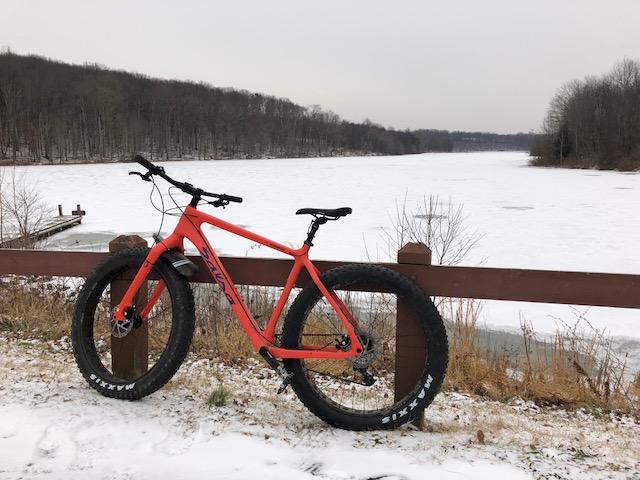 A bright orange fat bike is parked by a frozen lake, with a rustic wooden railing in the foreground. Snow covers the ground, and trees line the background under a cloudy sky. The lake is partially covered with ice, suggesting a winter setting. Black Hill Regional Park mountain bike trail.