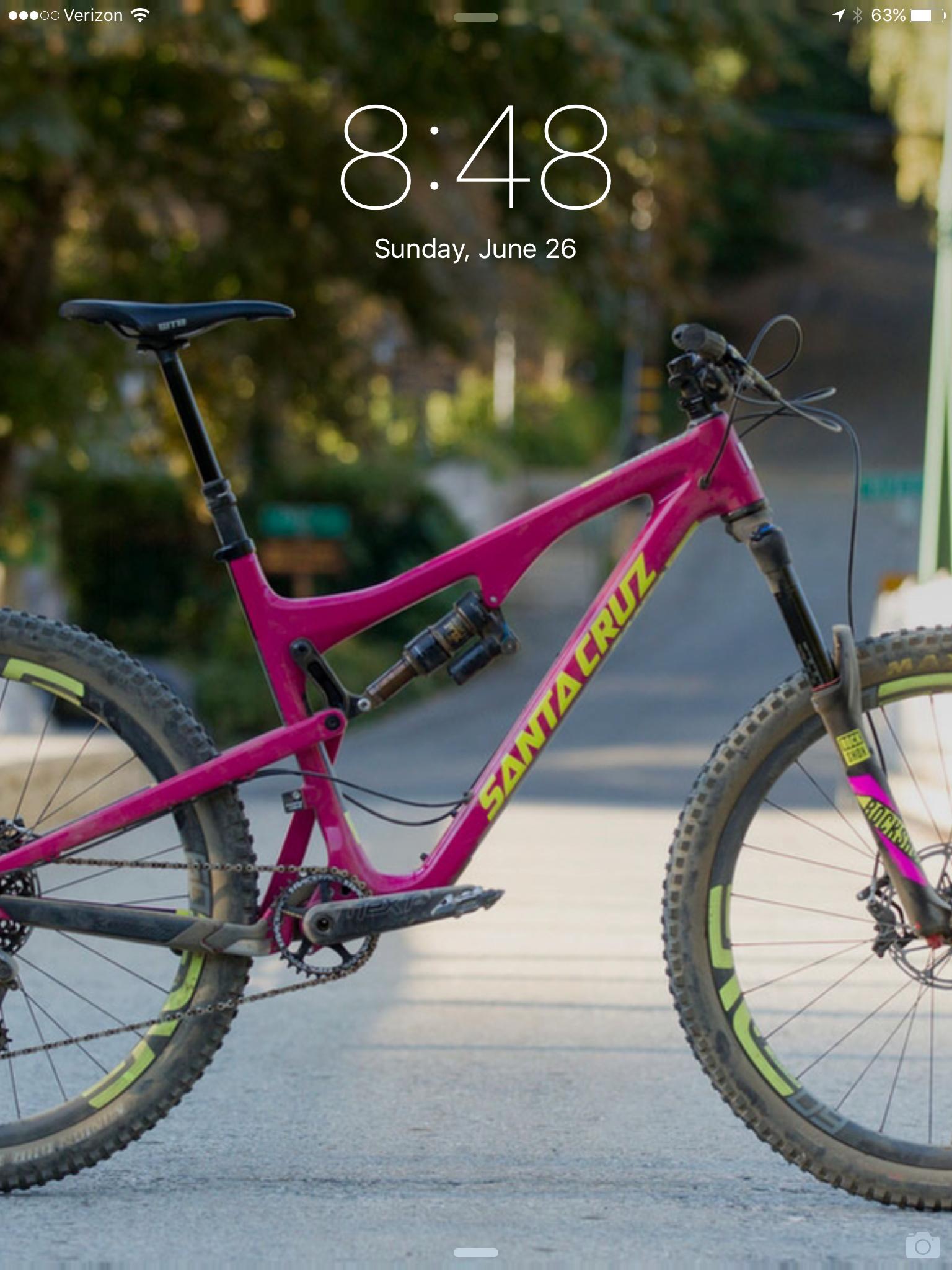 Santa Cruz Bronson C: A vibrant pink mountain bike with yellow accents, positioned on a paved path surrounded by trees. The bike features a full suspension system, a sleek frame, and thick tires designed for performance. The background is slightly blurred, highlighting the bike while showing a glimpse of a scenic outdoor setting. The time on the screen is 8:48 on Sunday, June 26.