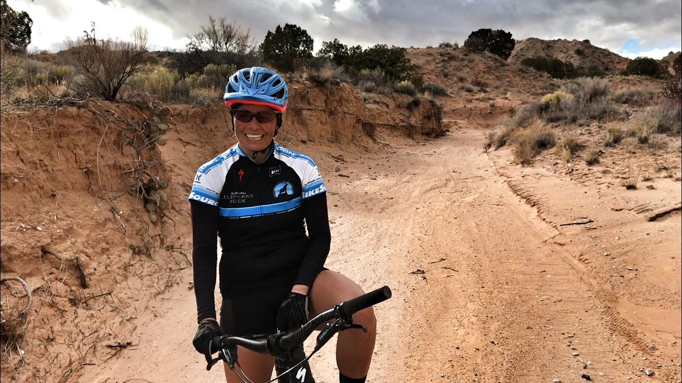 A mountain biker wearing a blue and black cycling jersey and a helmet is smiling, sitting on a bike on a sandy trail surrounded by rocky terrain and sparse vegetation, with a cloudy sky in the background. Mariposa Fat Bike Trails mountain bike trail.