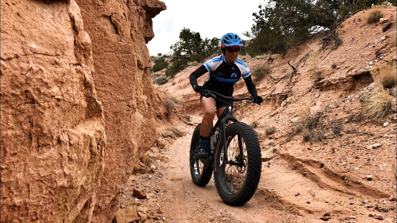A cyclist riding a fat bike on a narrow dirt trail surrounded by red rock formations and sparse vegetation, showcasing an outdoor adventurous scene. The cyclist is wearing a blue helmet and a short-sleeve jersey while navigating the terrain. Mariposa Fat Bike Trails mountain bike trail.