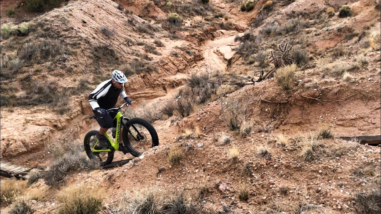 A mountain biker navigates a rocky, uneven trail, climbing up a steep incline in a desert landscape. The cyclist is wearing a helmet, gloves, and a jersey, riding a green mountain bike, with dry vegetation and rocky terrain surrounding them. Mariposa Fat Bike Trails mountain bike trail.