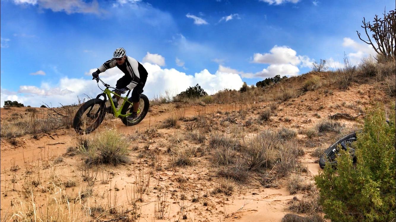 A person riding a fat bike down a sandy trail in a desert landscape, surrounded by sparse vegetation and under a partly cloudy sky. Mariposa Fat Bike Trails mountain bike trail.