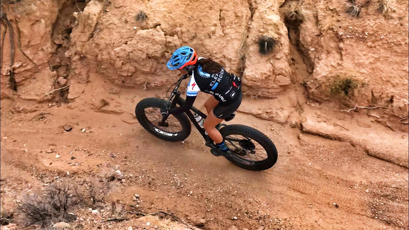 A cyclist riding a fat bike on a sandy, rugged trail surrounded by rocky terrain. The rider is wearing a blue helmet and a black cycling jersey with logos, focused on navigating the path. Mariposa Fat Bike Trails mountain bike trail.