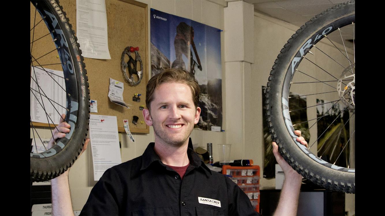 A smiling man in a black shirt holds up two mountain bike tires in a bicycle shop, with a bulletin board and bike-related posters in the background.