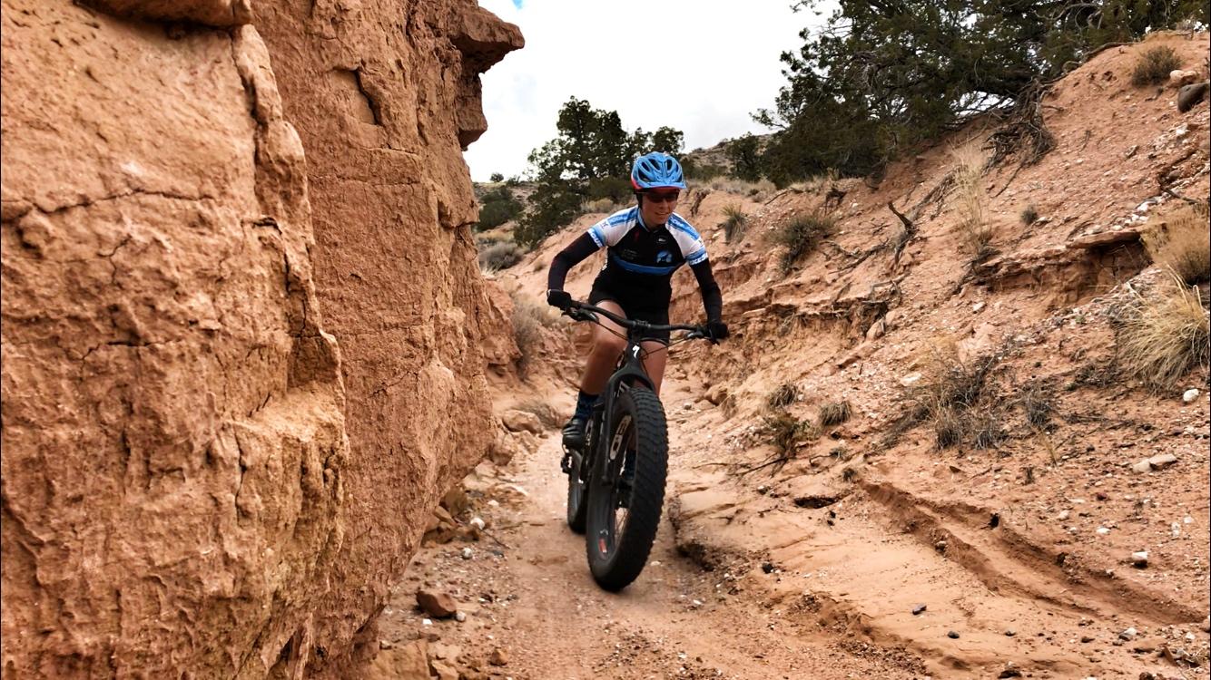 A cyclist riding a fat tire bike on a narrow dirt trail surrounded by rocky terrain and sparse vegetation. The rider is wearing a blue helmet and a cycling jersey, navigating through the rugged landscape. Mariposa Fat Bike Trails mountain bike trail.