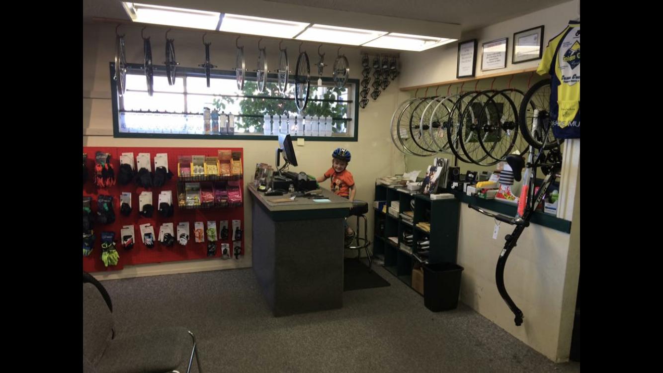 A cozy bike shop interior featuring a young child wearing a bicycle helmet sitting at a counter with a computer. The walls display various bicycle wheels and accessories for sale, including gloves and snacks, organized on shelves and pegboards. Natural light filters in through a window, creating a welcoming atmosphere.