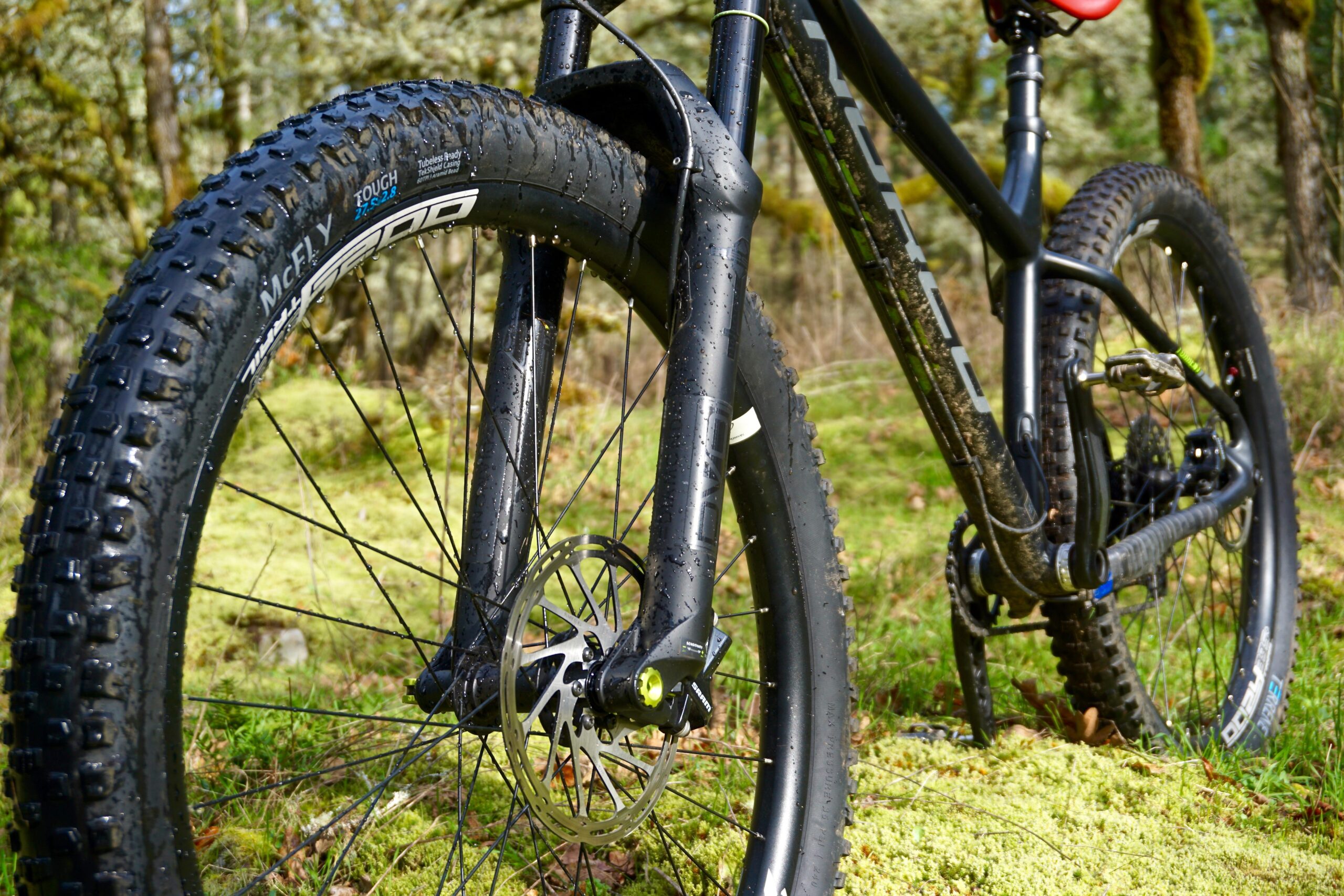 Terrene McFly: Close-up of a mountain bike front wheel and fork, showing a rugged tire with pronounced tread, a hydraulic disc brake, and droplets of water on the frame. The background features a mossy forest floor, indicative of a natural outdoor setting.