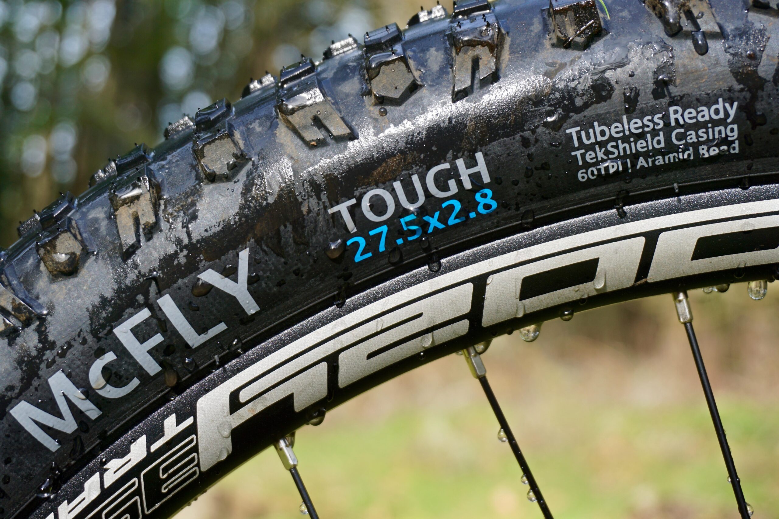Terrene McFly: Close-up image of a mountain bike tire with the brand name "McFLY" visible. The tire features a tread pattern designed for traction, labeled "TOUGH" with dimensions "27.5x2.8" in blue. It also displays information about being tubeless ready and TekShield casing technology. Water droplets are present on the tire, suggesting recent exposure to wet conditions. The background is blurred, emphasizing the tire.
