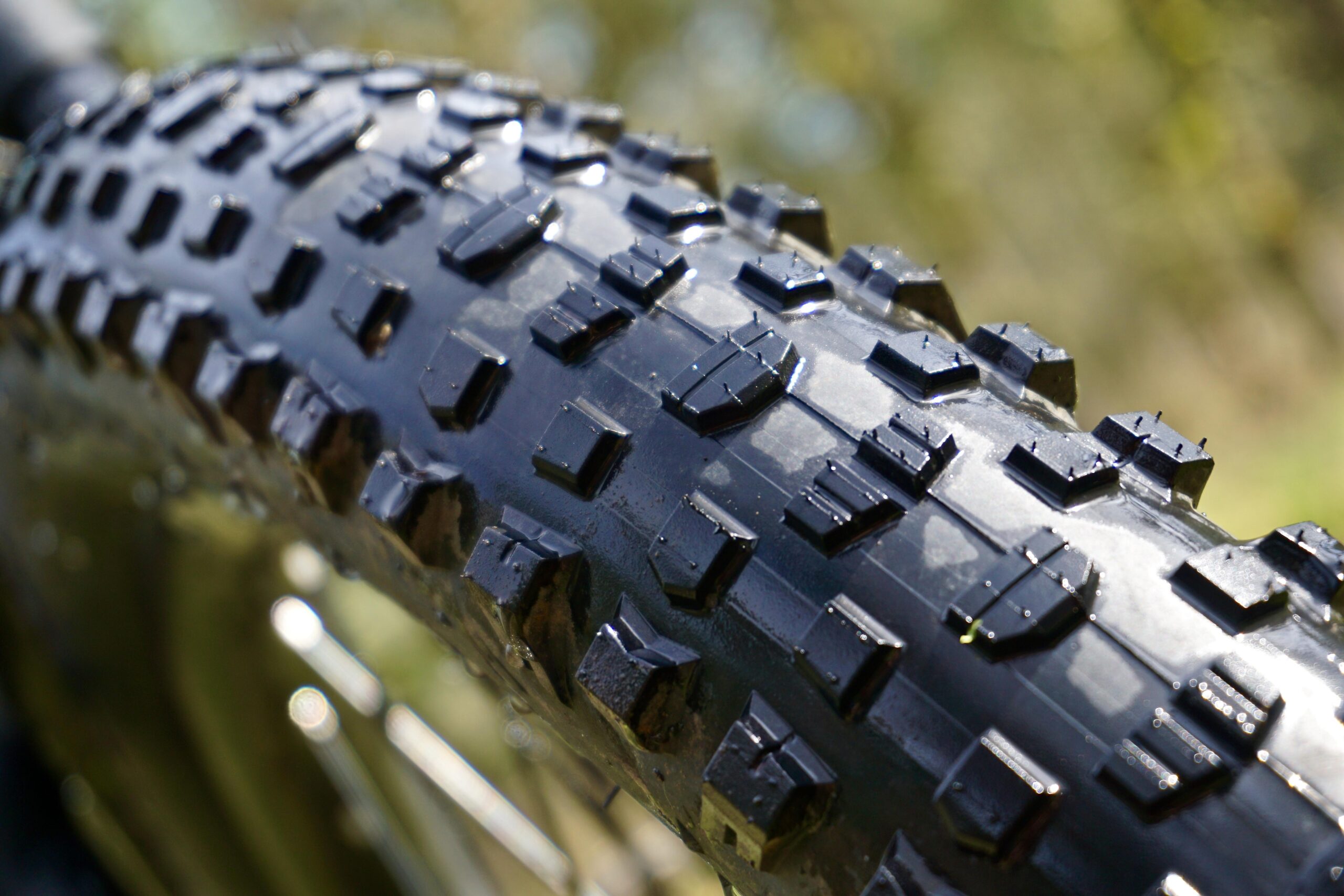 Terrene McFly: Close-up image of a bicycle tire showcasing its textured tread pattern, designed for enhanced grip and traction. The tire is wet, indicating recent exposure to water or mud, and the background features a blurred, natural setting.