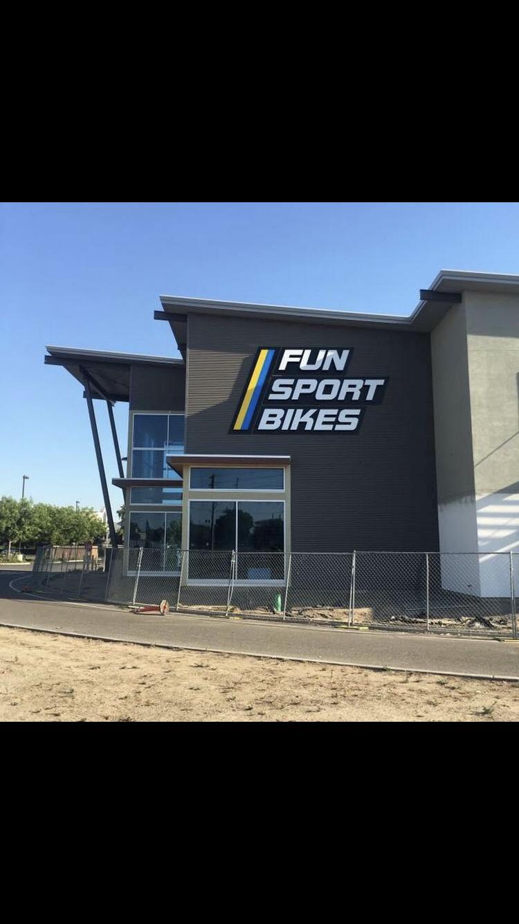 Image of a modern building featuring a prominent sign that reads "FUN SPORT BIKES." The structure has large glass windows and a sleek architectural design, with clear blue skies in the background and a fenced area in front.