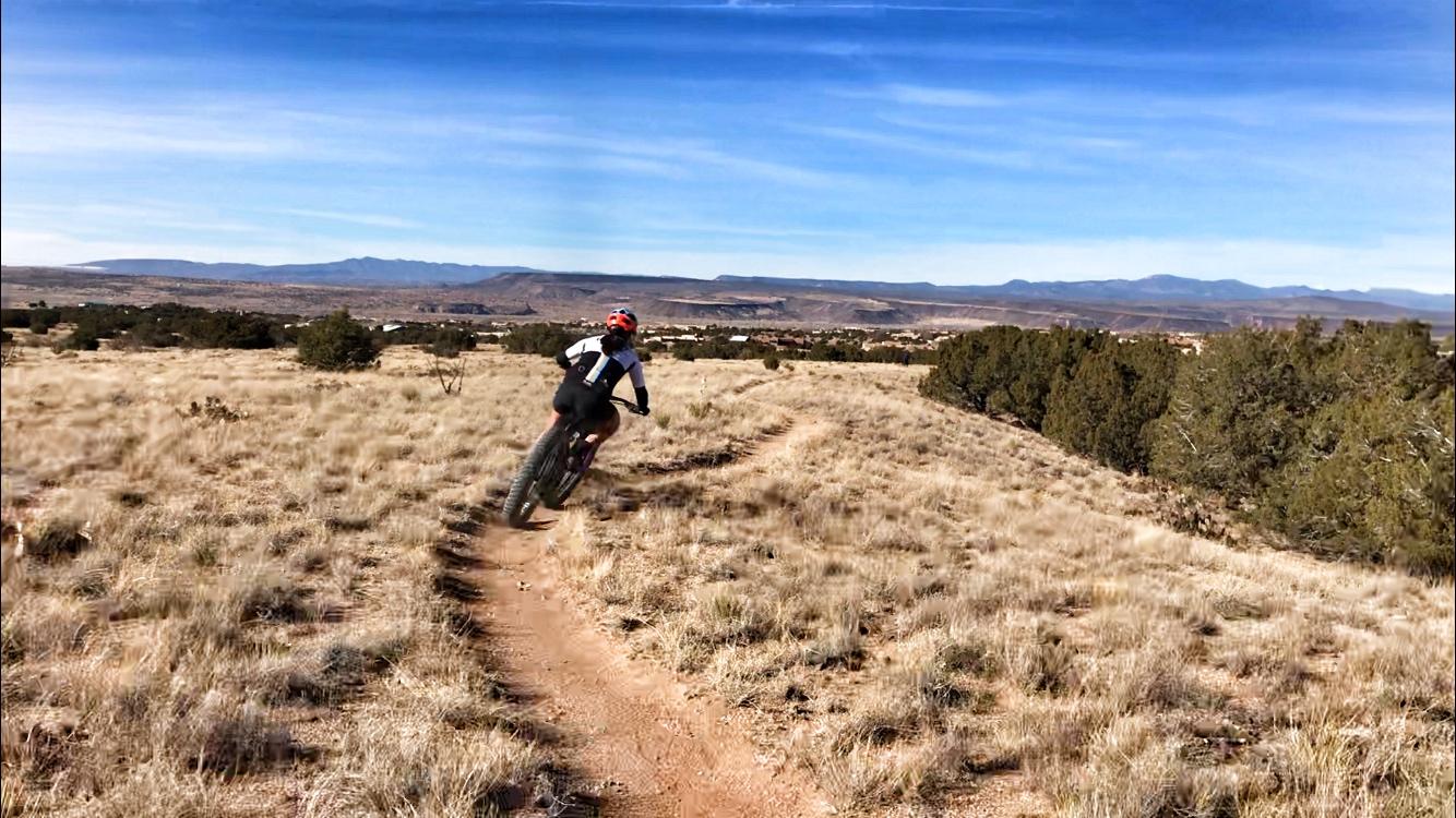 A cyclist riding a fat tire bike along a dirt trail in a grassy landscape, with distant mountains and a clear blue sky in the background. Placitas Trails mountain bike trail.
