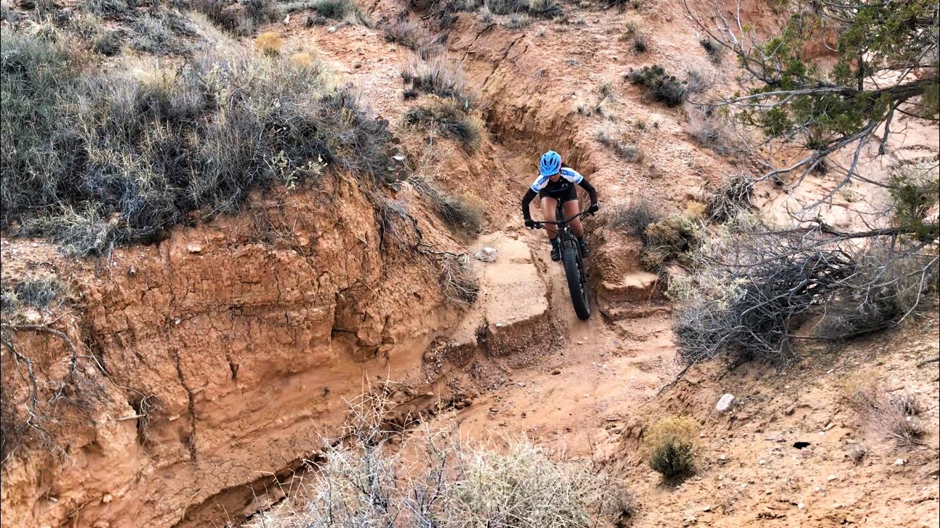A mountain biker navigating a rugged terrain with dry, reddish soil and sparse vegetation, set against a backdrop of steep, uneven slopes. The cyclist is wearing a blue helmet and a fitted cycling outfit, focused on maneuvering through a narrow path in the landscape. Mariposa Fat Bike Trails mountain bike trail.