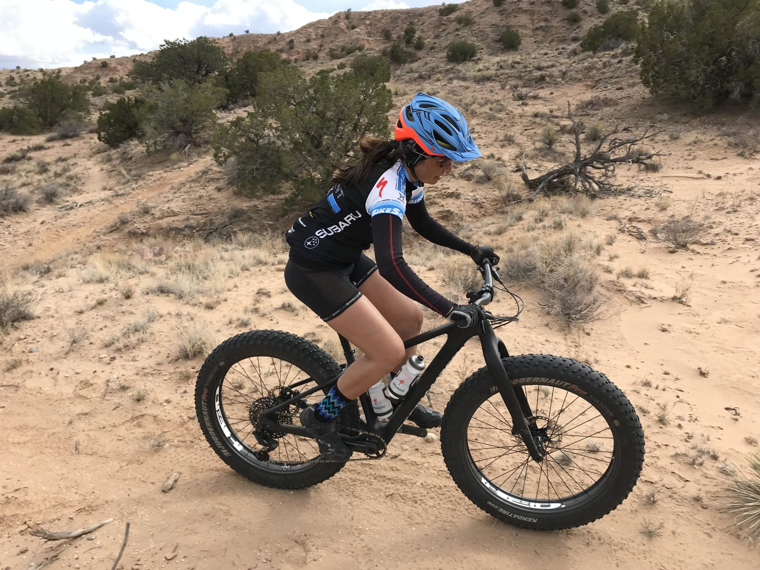 A young cyclist riding a fat tire bike on a sandy trail in a desert landscape, wearing a helmet and cycling attire. The background features sparse vegetation and hills under a partly cloudy sky. Mariposa Fat Bike Trails mountain bike trail.
