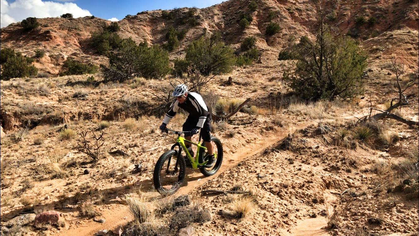 A person riding a bright green fat tire mountain bike on a dirt trail in a rugged, rocky landscape with sparse vegetation and hills in the background. Mariposa Fat Bike Trails mountain bike trail.