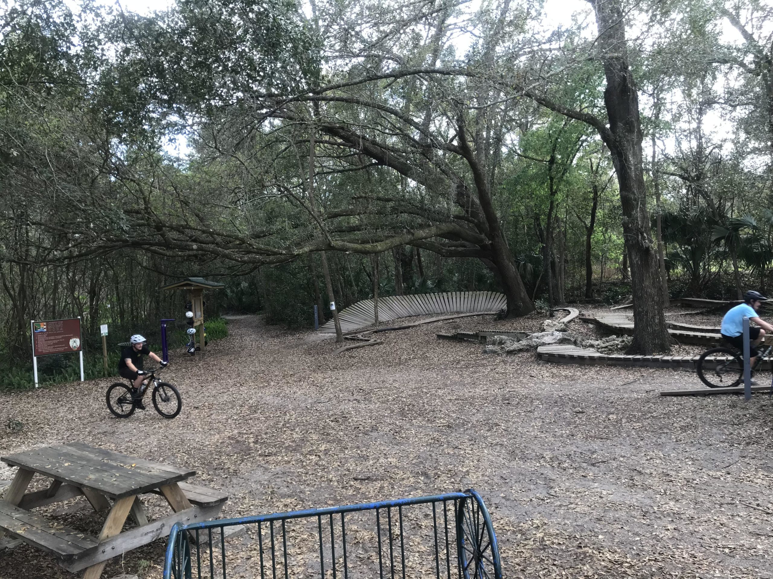 A scenic outdoor area featuring two mountain bikers navigating a trail surrounded by lush greenery. There is a picnic table in the foreground and various trail markers visible, along with a winding path through the trees. Leaves cover the ground, indicating a natural, wooded environment. Mount Dora Trail mountain bike trail.