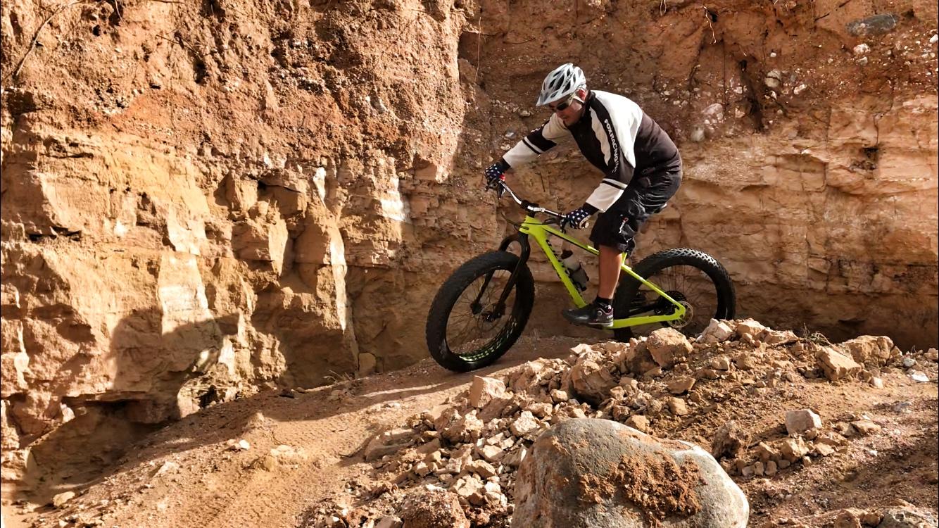 A mountain biker riding a bright green fat bike on rocky terrain, navigating a steep, uneven pathway with a dirt and rock background. The cyclist is wearing a helmet and protective gear, showcasing an action-packed moment of off-road biking. Mariposa Fat Bike Trails mountain bike trail.