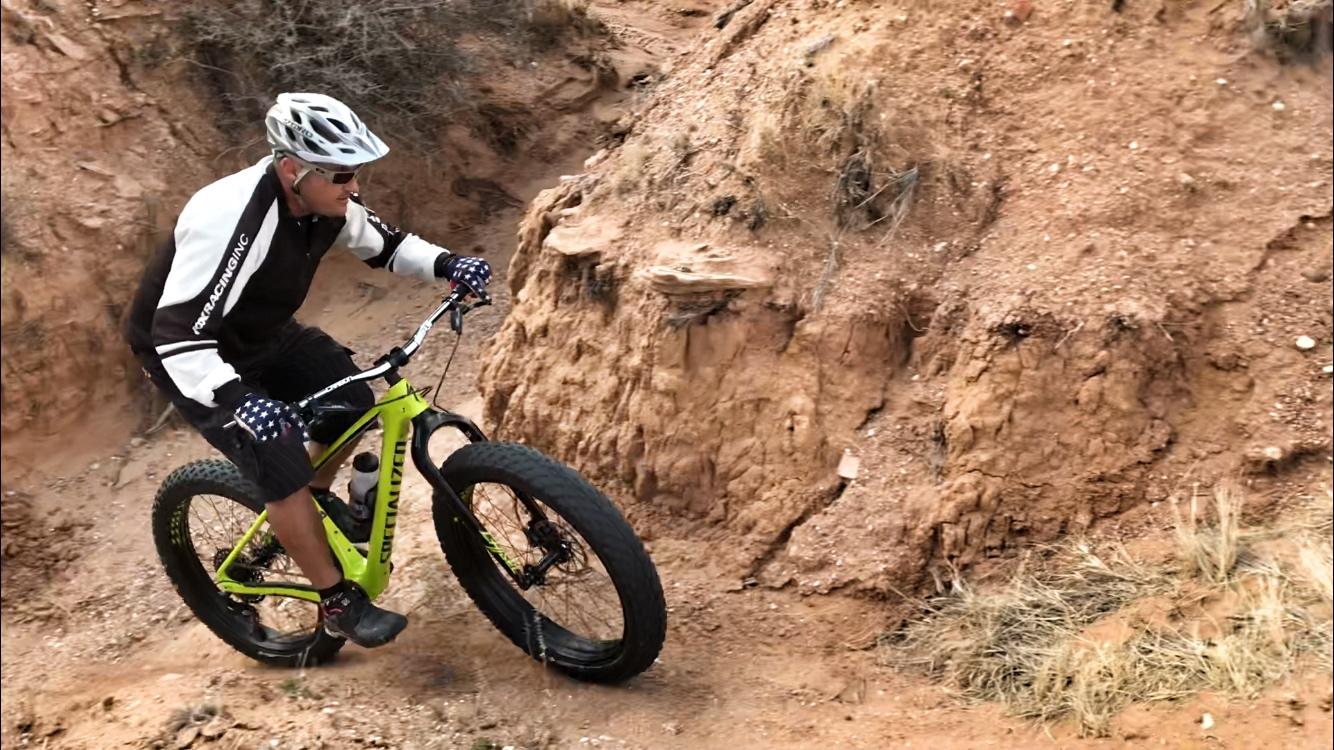 A cyclist navigating a rugged dirt trail on a bright green fat bike. The terrain features rocky and uneven ground, with steep slopes on either side. The rider is dressed in a black and white cycling jersey and gloves, wearing a helmet for safety. Mariposa Fat Bike Trails mountain bike trail.