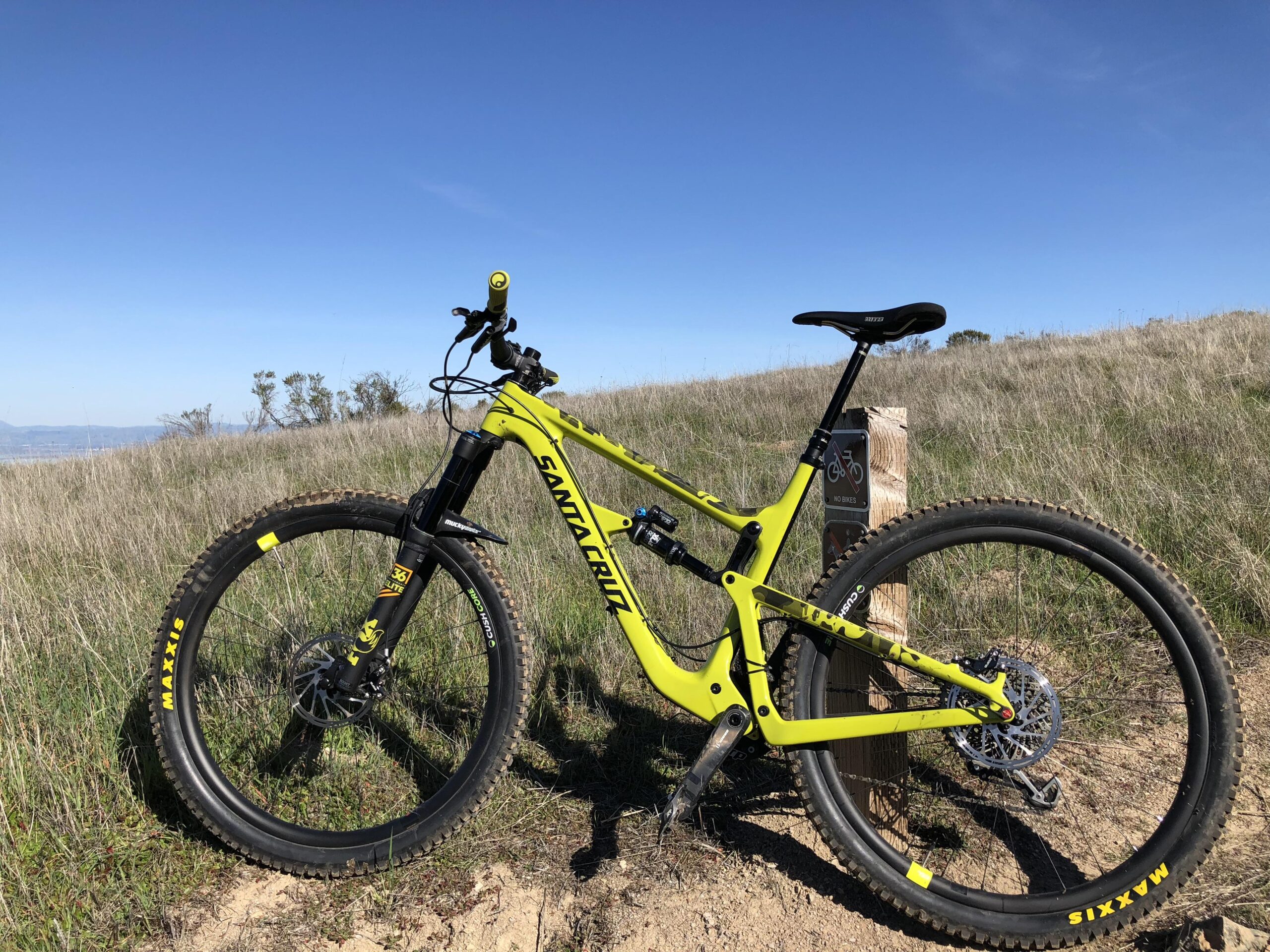 Santa Cruz Hightower LT: A bright yellow mountain bike parked on a grassy hill, with a clear blue sky in the background. The bike features large tires marked with "Maxxis" and a prominent suspension system, with a wooden post nearby indicating a "no bikes" sign.