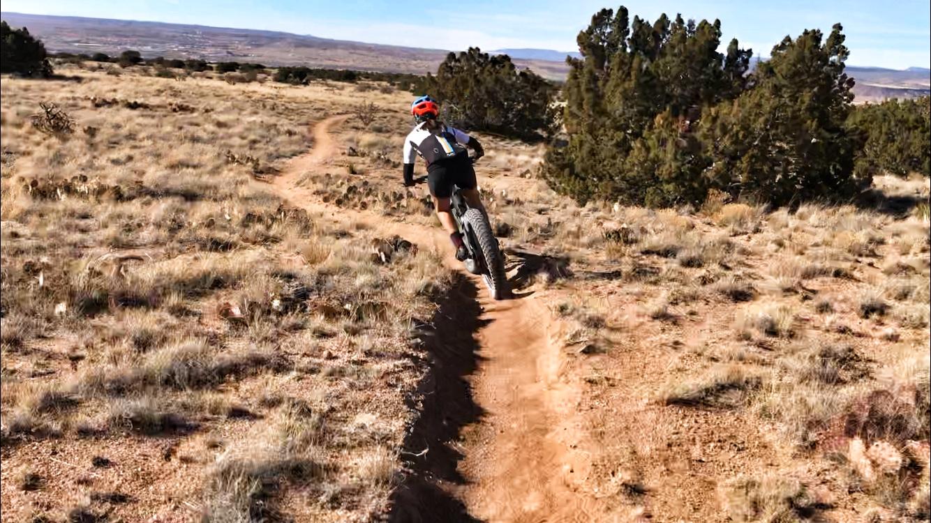 A mountain biker rides along a dirt trail in a rugged, grassy landscape with scattered trees and hills in the background. The cyclist is wearing a helmet and biking gear, showcasing a dynamic moment as they navigate a curved path. The scene captures the essence of outdoor adventure and sports. Placitas Trails mountain bike trail.