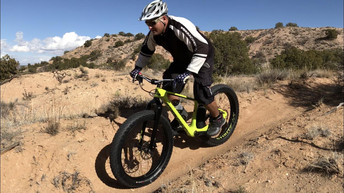 A person riding a bright yellow fat bike on a sandy trail, surrounded by sparse vegetation and hills under a clear blue sky. The rider is wearing a helmet and protective gear, focused on navigating the terrain. Mariposa Fat Bike Trails mountain bike trail.
