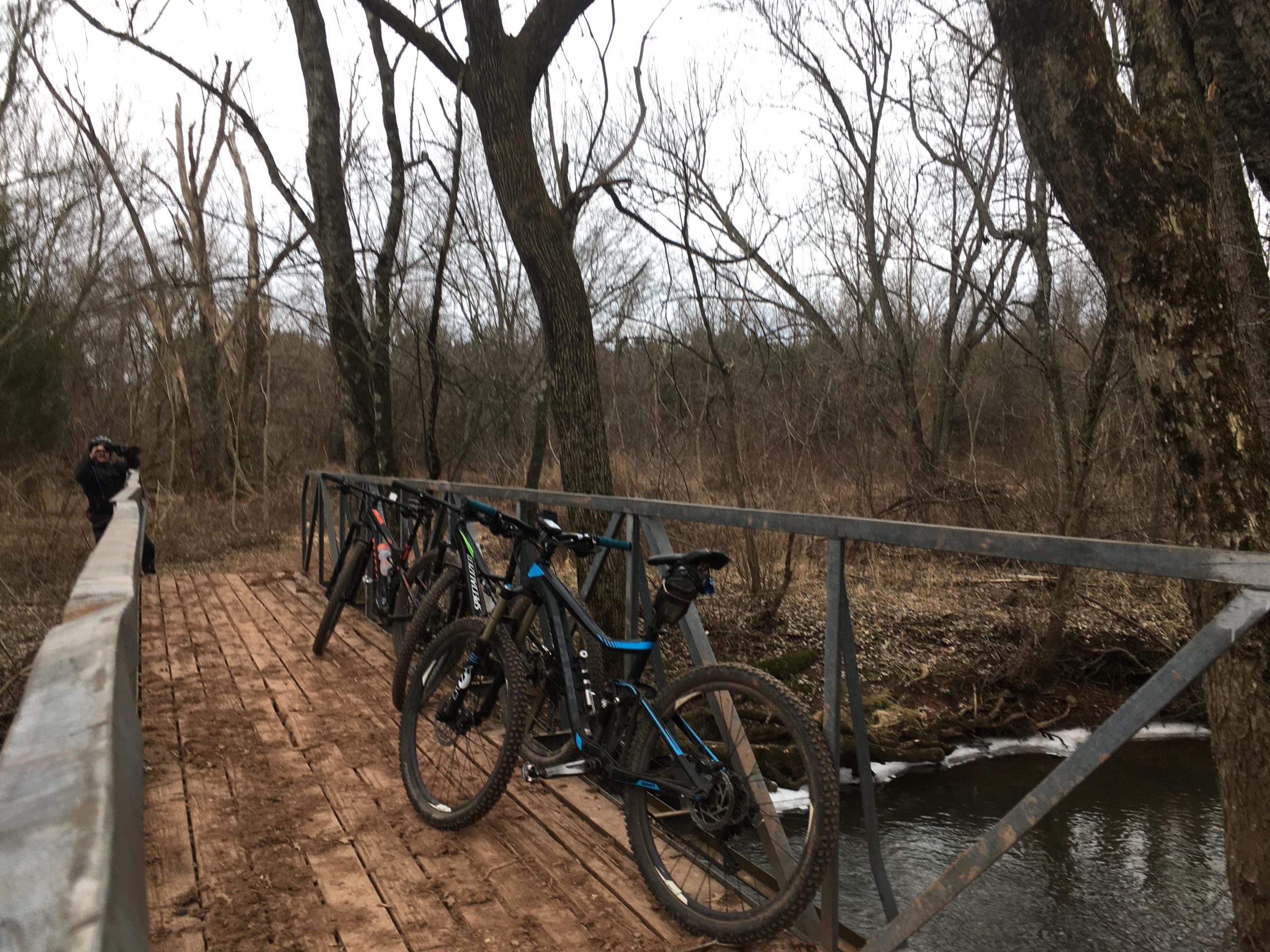 A wooden bridge over a creek surrounded by bare trees and brush, with several mountain bikes parked on the bridge. A person in the background is taking a photo. The scene conveys a tranquil outdoor setting suitable for cycling and exploration. Six Mile Run mountain bike trail.