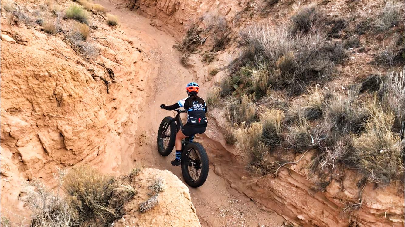A cyclist riding a fat tire bike along a sandy, winding trail in a rocky landscape, surrounded by sparse vegetation and rugged terrain. The cyclist wears a helmet and a jersey, focusing on navigating the path ahead. Mariposa Fat Bike Trails mountain bike trail.