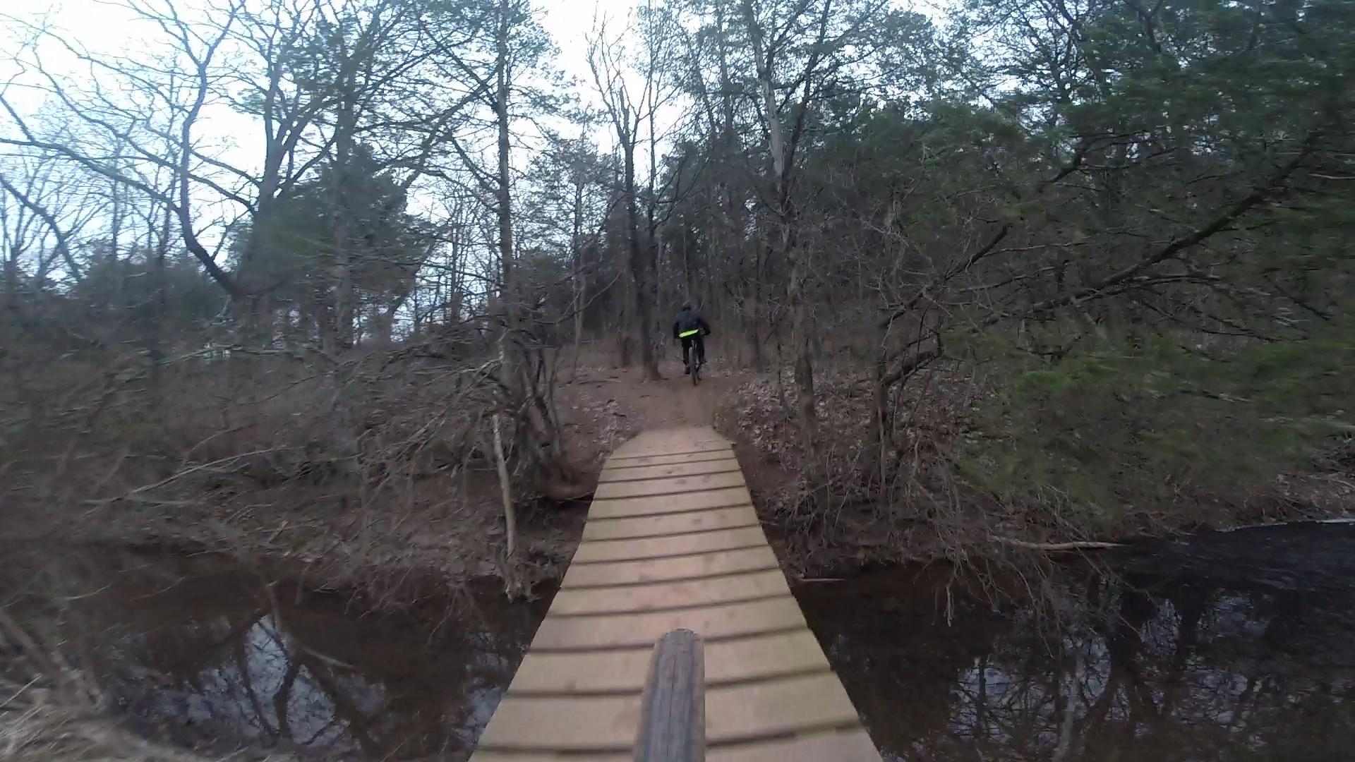 A mountain biker riding across a wooden bridge over a small stream, surrounded by bare trees and a natural landscape. The scene is set in a forested area on a cloudy day. Six Mile Run mountain bike trail.