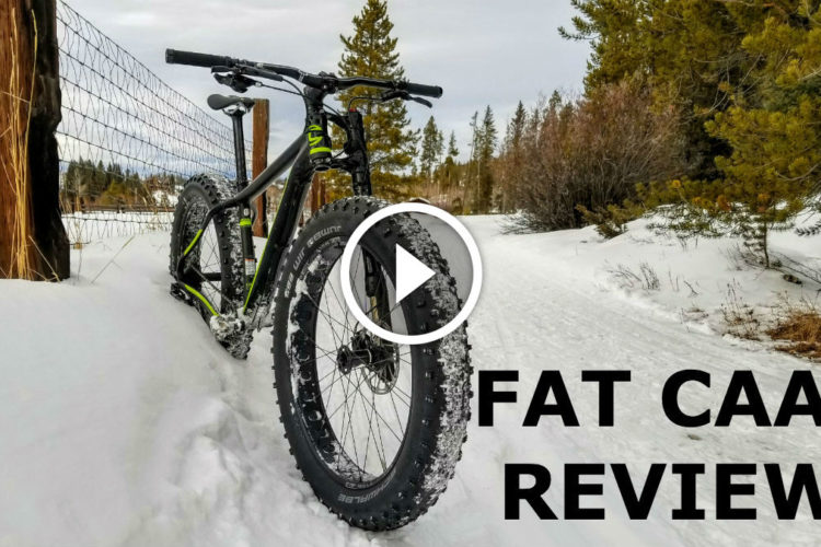 A black and green fat bike parked on a snowy trail, with a wooden fence in the background and trees lining the path. A play button overlay indicates a video review titled "Fat CAAD Review."