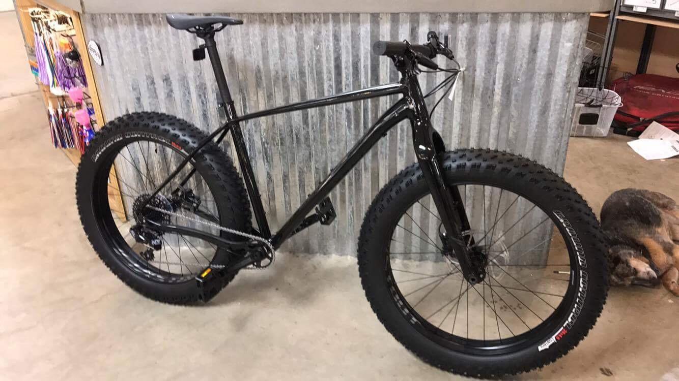 Specialized Fatboy: A black fat bike with wide tires stands in a indoor space, leaning against a corrugated metal wall. Various colorful items are displayed in the background on wooden shelves, and a small animal is resting nearby on the floor.